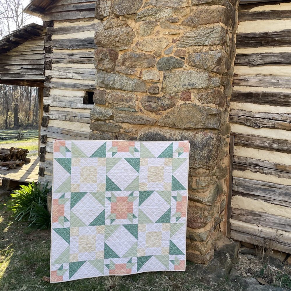 Handcrafted quilt with pastel patterns displayed outside a rustic stone and wood cabin by Heart Lake Quilts.