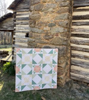 Handcrafted quilt with pastel patterns displayed outside a rustic stone and wood cabin by Heart Lake Quilts.