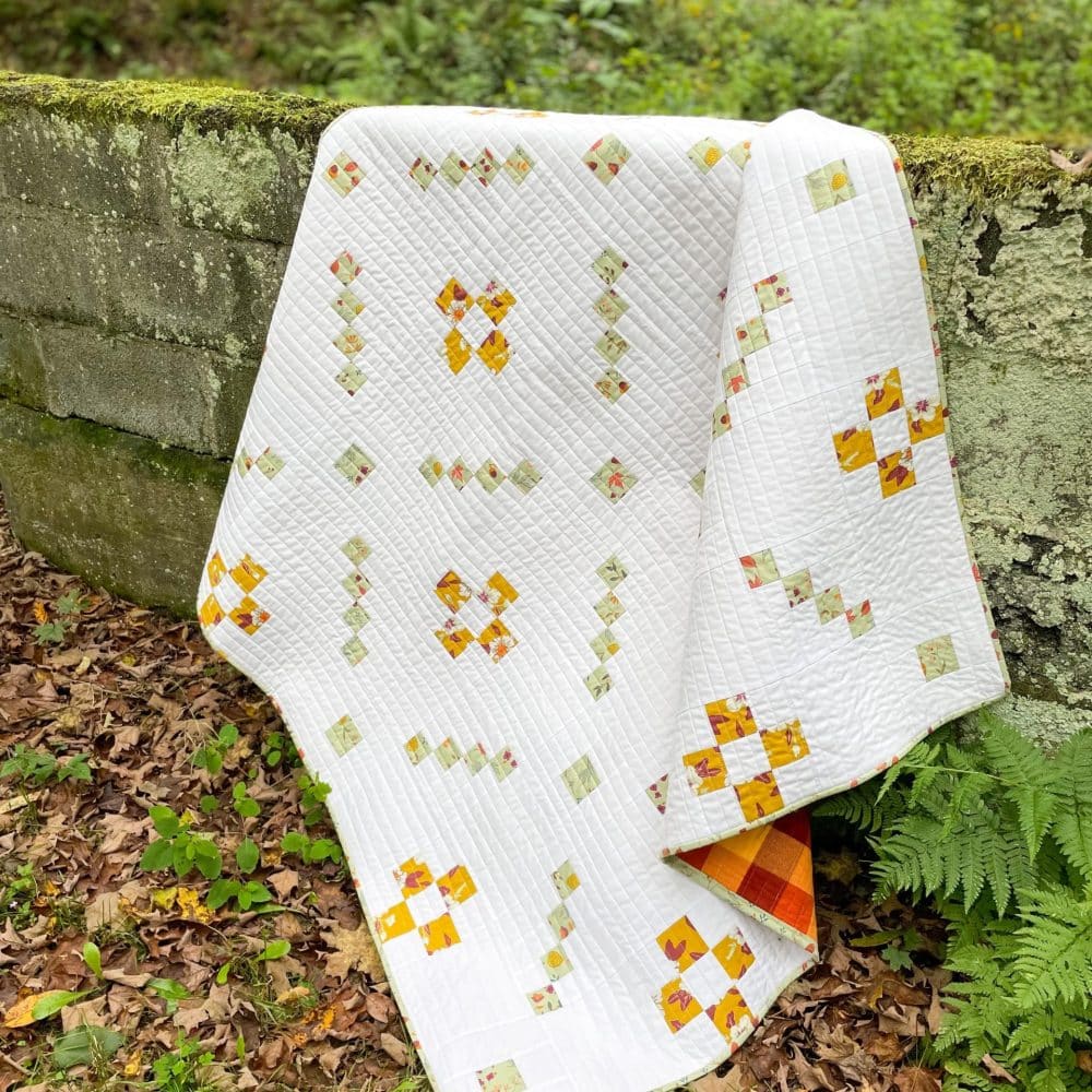 Handmade white quilt with yellow and green patterns draped over mossy wall in forest - Heart Lake Quilts design