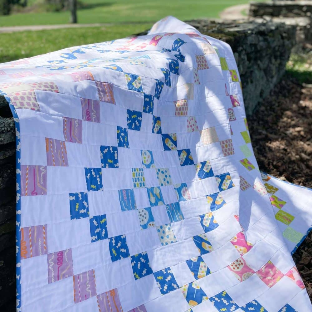 Colorful handmade quilt draped over stone wall outdoors by Heart Lake Quilts, featuring bright patchwork patterns.