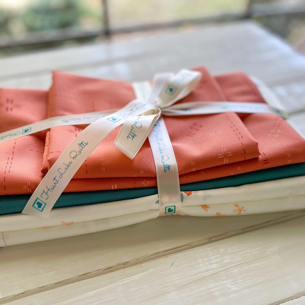 Heart Lake Quilts fabric bundle with orange, green, and white patterned fabrics tied with branded ribbon.