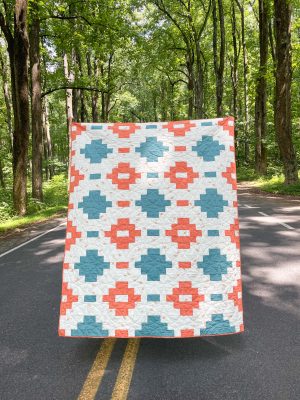 Handcrafted quilt with geometric pattern in teal and coral by Heart Lake Quilts, displayed in a scenic forest setting.