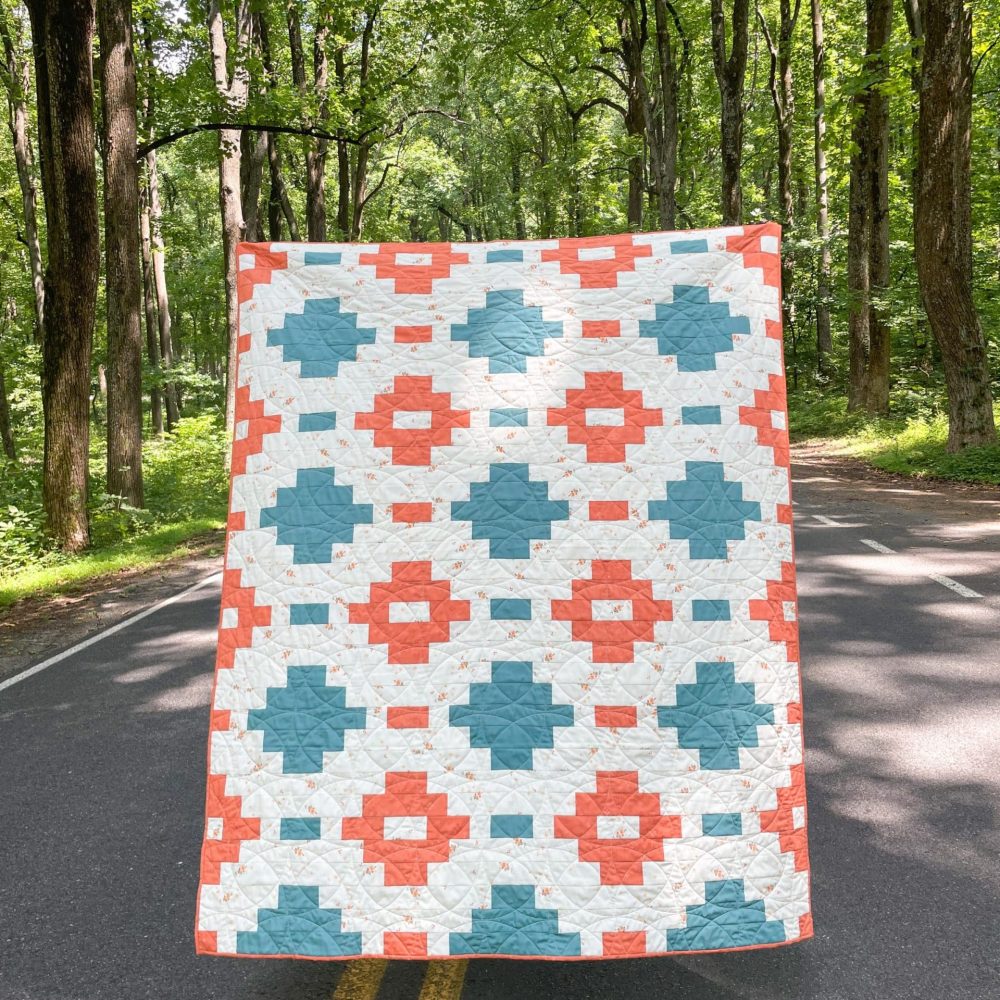 Handcrafted quilt with geometric pattern in teal and coral by Heart Lake Quilts, displayed in a scenic forest setting.